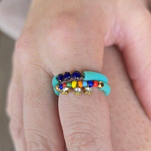 Puede incluir: Dos anillos turquesa con detalles de cuentas de colores. Un anillo presenta una fila de cuentas azules, mientras que el otro tiene un arcoíris de cuentas y adornos dorados con piedras transparentes. Joyería.