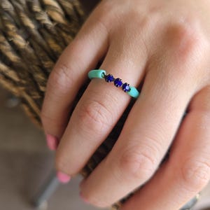 May include: A close-up of a turquoise ring with three dark blue gemstones. The ring is on a finger, and the background is blurred.
