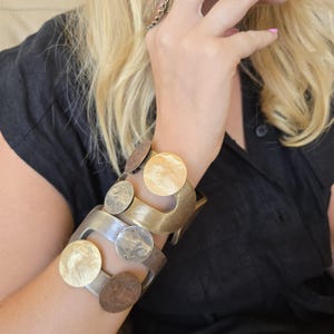 Könnte beinhalten: Zwei breite Manschettenarmbänder aus Metall am Handgelenk. Jedes Armband hat eine gebürstete Oberfläche und ist mit mehreren kreisförmigen Metallscheiben in verschiedenen Gold-, Bronze- und Silbertönen verziert, was für eine Textur sorgt.