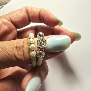 Op de afbeelding: Twee ringen gedragen aan een vinger. Eén ring heeft een rij kleine witte parels gescheiden door zilverkleurige tussenstukjes. De tweede ring is een zilverkleurige band bezet met drie heldere, gefacetteerde stenen in een verticale lijn.