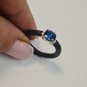 May include: A black ring with a vibrant blue gemstone set in a silver-colored metal. The gemstone is square-cut and catches the light, creating a sparkling effect. The ring is held against a white background.