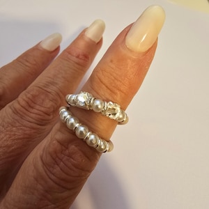 May include: Two silver rings are displayed on a finger. One ring features a row of small pearls, while the other has a central clear gemstone flanked by smaller pearls. The rings are set against a neutral background.