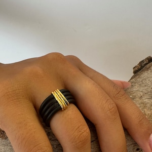 Puede incluir: Un anillo de goma negro con una banda dorada envuelta alrededor. El anillo se lleva en un dedo.