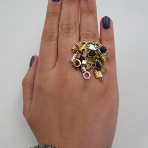 May include: A gold ring with a cluster of charms, including small chains, beads, and geometric shapes. The ring is worn on a hand with dark blue nail polish.