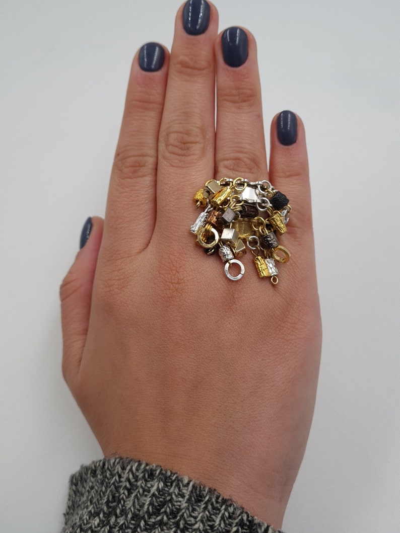 May include: A gold ring with a cluster of charms, including small chains, beads, and geometric shapes. The ring is worn on a hand with dark blue nail polish.