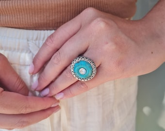 Puede incluir: Un anillo de plata con una piedra turquesa engastada en un diseño circular. El anillo tiene un borde decorativo alrededor de la piedra.