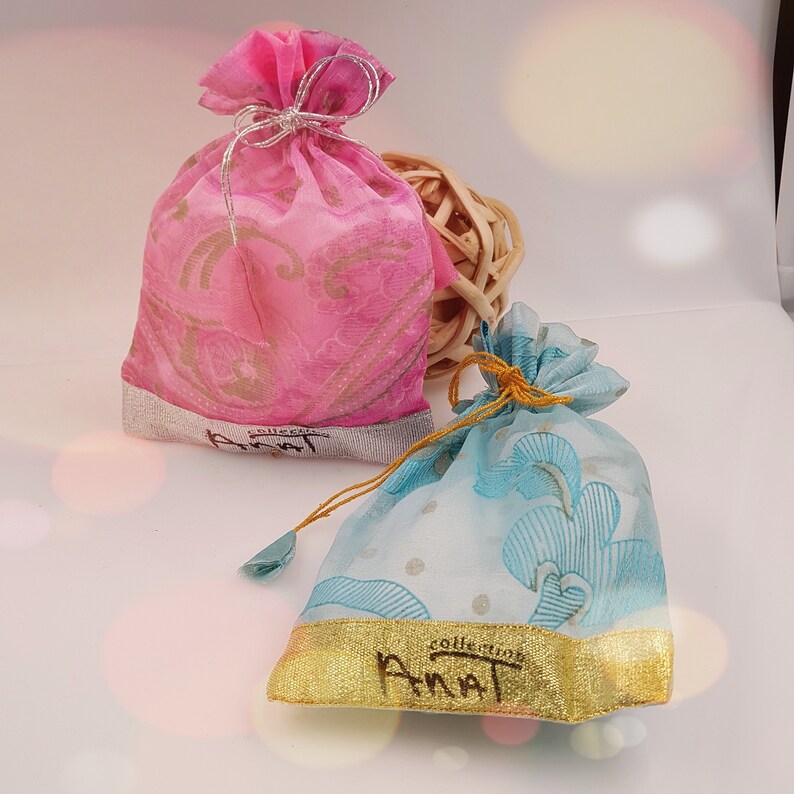 May include: Two small, decorative drawstring bags made of sheer fabric. The bag on the left is pink with a gold floral pattern and the bag on the right is light blue with a white and gold floral pattern. Both bags have a gold trim and the word "Annat" printed on the bottom.