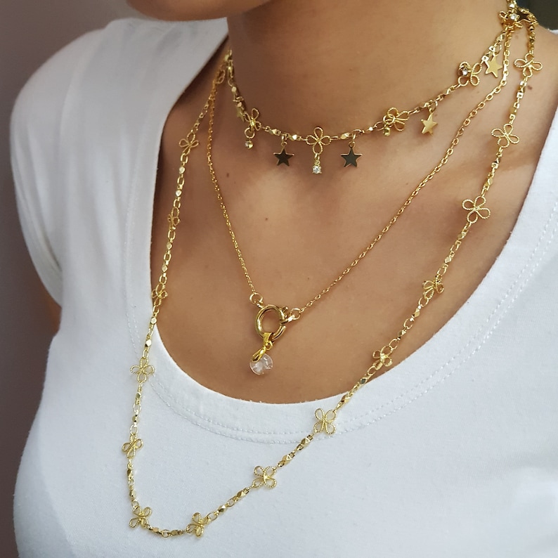 May include: Three gold necklaces with delicate flower and star charms. The top necklace has a star charm with a small crystal in the center. The bottom necklace has a crystal pendant with a gold clasp.