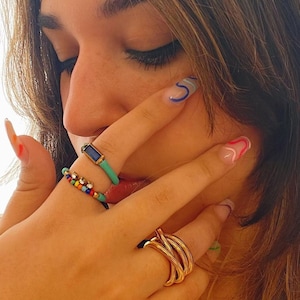 May include: A woman's hand wearing a variety of rings, including a blue and silver ring, a multi-colored beaded ring, and a gold ring with a twisted design. The rings are on her index, middle, and ring fingers.