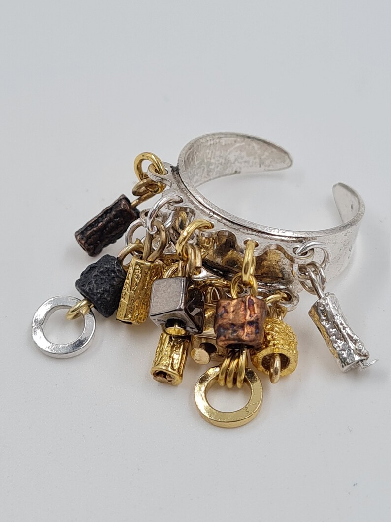 May include: A silver ring with multiple charms hanging from it. The charms are made of different metals, including gold, silver, and copper. The charms are in various shapes, including squares, circles, and cylinders.
