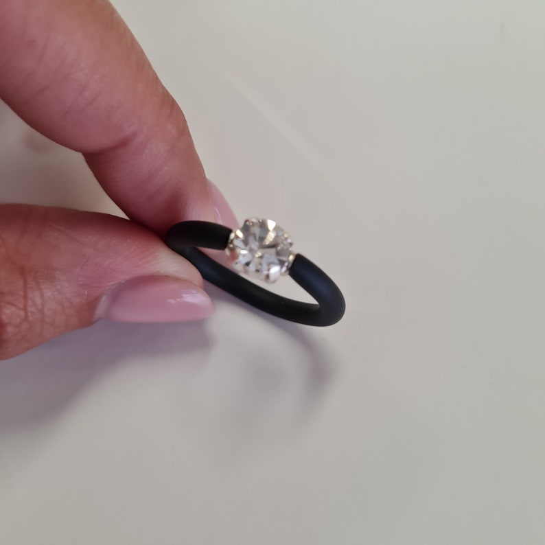 K&ouml;nnte beinhalten: Ein schwarzer Silikonring mit einem gro&szlig;en, klaren, runden Edelstein. Der Edelstein ist in einem silberfarbenen Metall gefasst. Der Ring hat ein schlichtes, modernes Design.