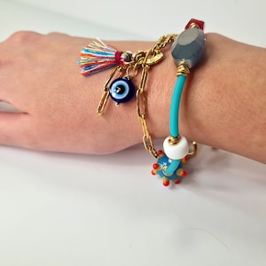 Puede incluir: Un brazalete colorido con un amuleto de ojo azul, un amuleto de cuentas turquesa, una cuenta blanca y un amuleto de piedra gris. El brazalete está hecho de cadena dorada y cuerda turquesa.
