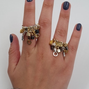 May include: Two gold and silver charm rings with dangling charms. The charms are a mix of gold, silver, and black metal. The rings are worn on the index and middle fingers of a hand.
