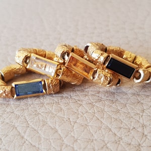 Könnte beinhalten: Vier goldene Ringe mit verschiedenen Edelsteinen. Die Ringe sind übereinander gestapelt. Die Edelsteine sind ein klarer Stein, ein orangefarbener Stein, ein schwarzer Stein und ein blauer Stein.