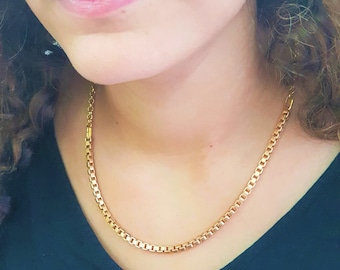 May include: A gold chain necklace with a box chain style. The chain is worn by a person with a black shirt.