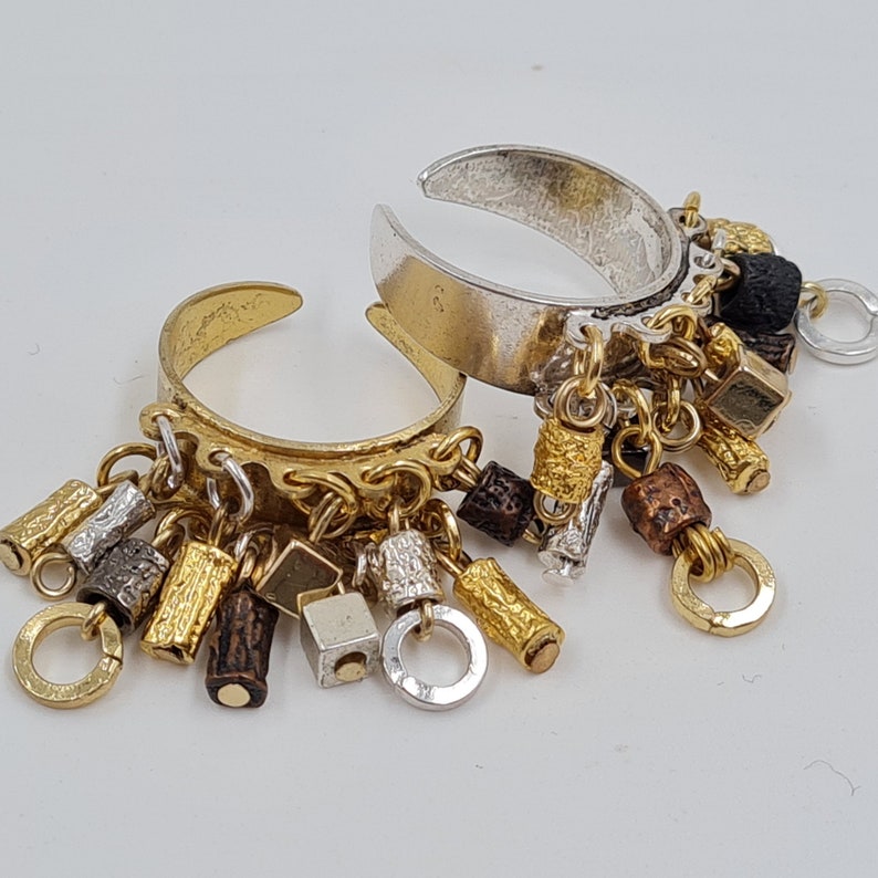 May include: Two silver and gold rings with dangling charms. The charms are made of various metals and include small squares, cylinders, and beads. The rings are a unique and stylish accessory.