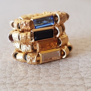 Könnte beinhalten: Goldring mit vier übereinanderliegenden Bändern, jedes mit einem rechteckigen Edelstein in einer anderen Farbe. Die Edelsteine sind blau, schwarz, orange und klar.