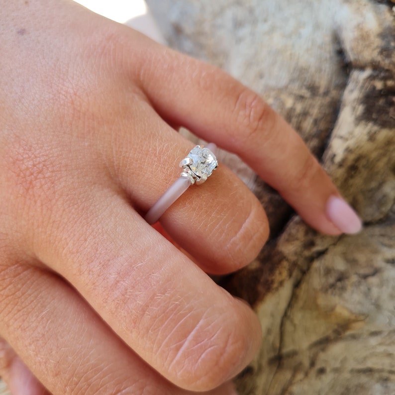 K&ouml;nnte beinhalten: Ein silberner Ring mit einem gro&szlig;en, klaren Edelstein. Der Ring hat ein hellrosa Band und wird an einem Finger getragen. Der Hintergrund ist ein verschwommenes Muster aus Braun und Beige.