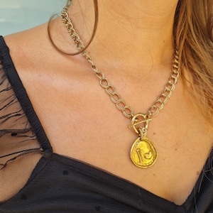 May include: A gold chain necklace with a large, round pendant featuring a stylised design. The pendant is attached to the chain with a toggle clasp.