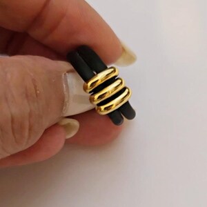 Könnte beinhalten: Ein schwarzer Ring mit drei goldfarbenen Bändern, die darum gewickelt sind. Der Ring wird von einer Hand vor einem weißen Hintergrund gehalten. Die goldenen Bänder haben ein glänzendes Finish.