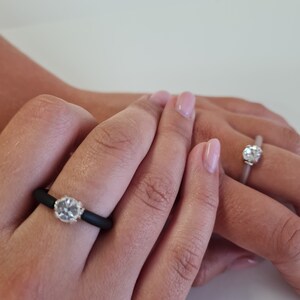 K&ouml;nnte beinhalten: Zwei Ringe mit einem klaren, runden Edelstein. Ein Ring hat ein schwarzes Band, der andere ein hellgraues Band. Die Ringe befinden sich an den Fingern einer Person, vor einem neutralen Hintergrund.