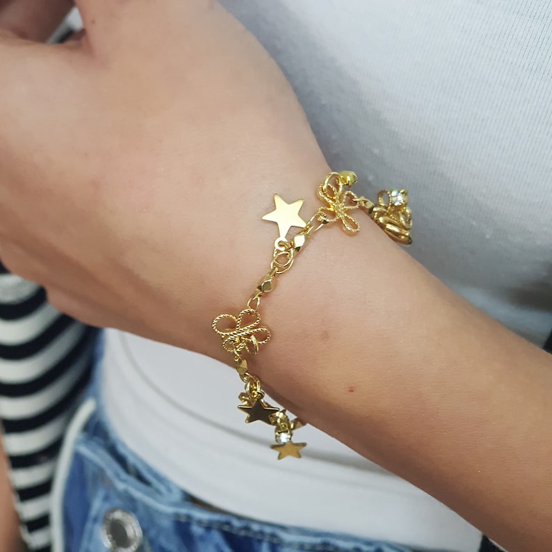 May include: A gold charm bracelet with star and bow charms. The bracelet is on a person's wrist.