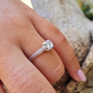 K&ouml;nnte beinhalten: Ein silberner Ring mit einem gro&szlig;en, klaren, rund geschliffenen Edelstein. Der Ring ist auf einem d&uuml;nnen, hellfarbigen Band gefasst. Der Edelstein wird von vier Krappen gehalten. Der Ring befindet sich an einem Finger.