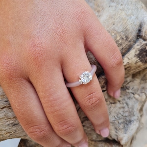 K&ouml;nnte beinhalten: Ein hellvioletter Silikonring mit einem gro&szlig;en, klaren, rund geschliffenen Edelstein und zwei kleineren Akzentsteinen. Der Ring befindet sich an einem Finger, vor einem nat&uuml;rlichen Holzuntergrund.