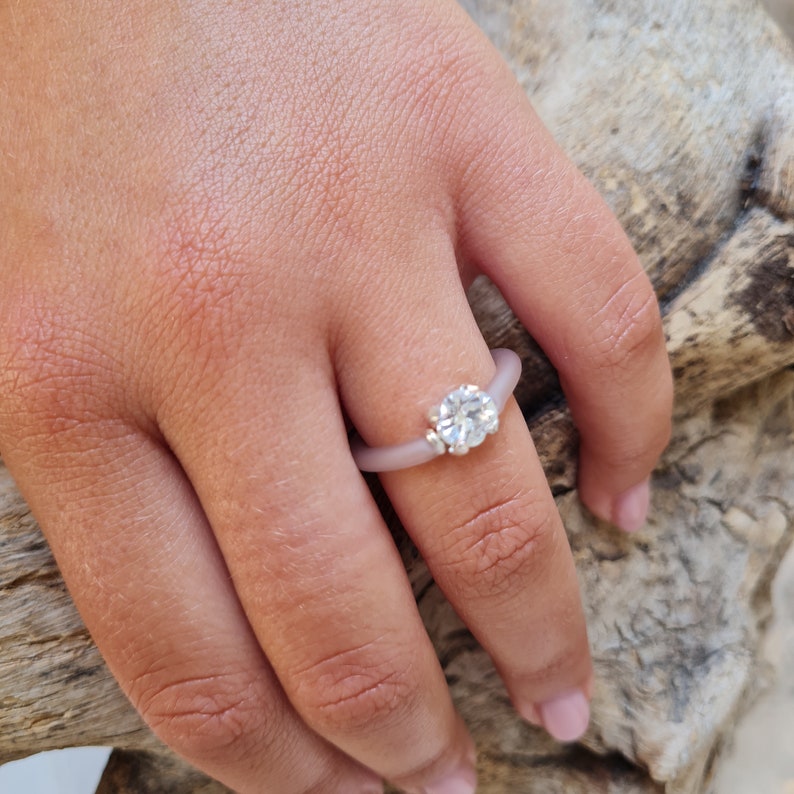 K&ouml;nnte beinhalten: Ein hellvioletter Silikonring mit einem gro&szlig;en, klaren, rund geschliffenen Edelstein und zwei kleineren Akzentsteinen. Der Ring befindet sich an einem Finger, vor einem nat&uuml;rlichen Holzuntergrund.
