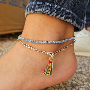 Puede incluir: Se muestran dos tobilleras. La tobillera superior es una fina hebra de piedras preciosas azul claro. La tobillera inferior es una cadena plateada con un pequeño colgante de borla de colores. Las tobilleras se llevan en un tobillo.