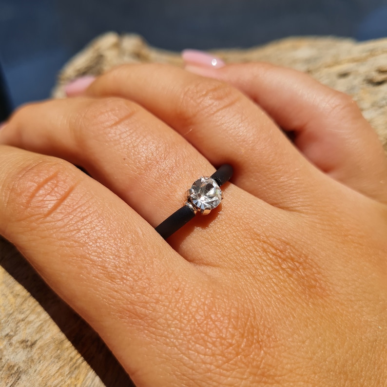 K&ouml;nnte beinhalten: Ein schwarzer Ring mit einem klaren, runden Edelstein, der in einem silberfarbenen Metall gefasst ist. Der Ring befindet sich an einem Finger, mit einem nat&uuml;rlichen Holzhintergrund.