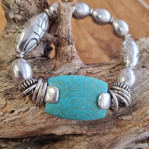 Op de afbeelding: Zilveren armband met een turquoise steen in het midden. De armband heeft een mix van zilveren kralen, waaronder ovale en bladvormige elementen, en is geplaatst tegen een natuurlijke houten achtergrond. De turquoise steen is rechthoekig met een getextureerd oppervlak.