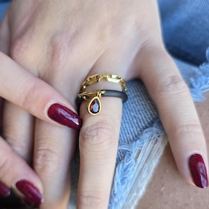 Op de afbeelding: Een gouden kettingring en een zwarte bandring met een druppelvormige rode edelsteenhanger. De ringen worden gedragen aan een vinger met donkerrode nagellak.
