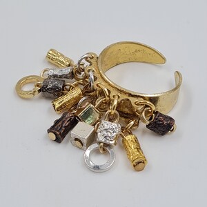 May include: A gold ring with multiple charms hanging from it. The charms are made of different metals and have various shapes, including squares, cylinders, and circles. Some of the charms are textured, while others are smooth.