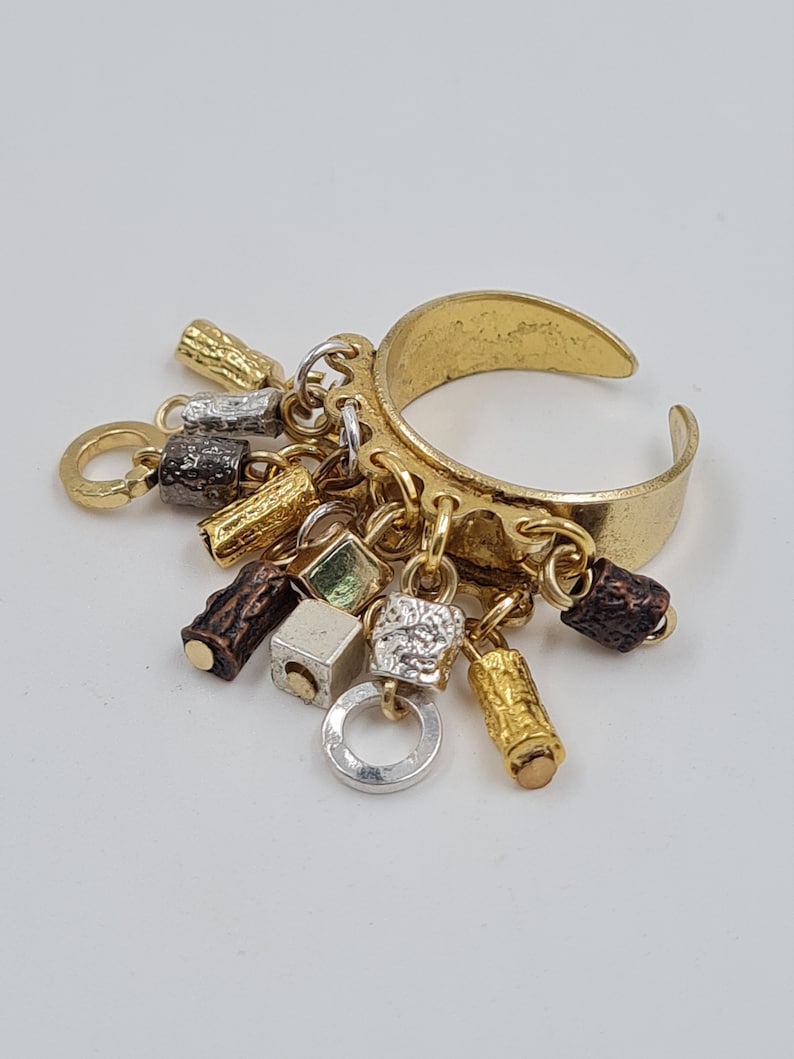 May include: A gold ring with multiple charms hanging from it. The charms are made of different metals and have various shapes, including squares, cylinders, and circles. Some of the charms are textured, while others are smooth.