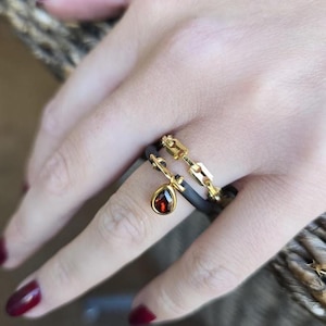 May include: A hand wearing two gold rings. One ring features a chain link design, and the other has a teardrop-shaped, red gemstone pendant. The rings are on a black band.