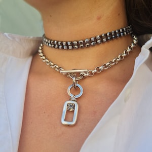 Könnte beinhalten: Eine zweireihige Halskette mit dunkelgrauen Kristallen und eine silberne Halskette mit einem rechteckigen Anhänger. Der Anhänger hat einen einzelnen Kristall in der Mitte. Der Schmuck wird über einem weißen Hemd getragen.