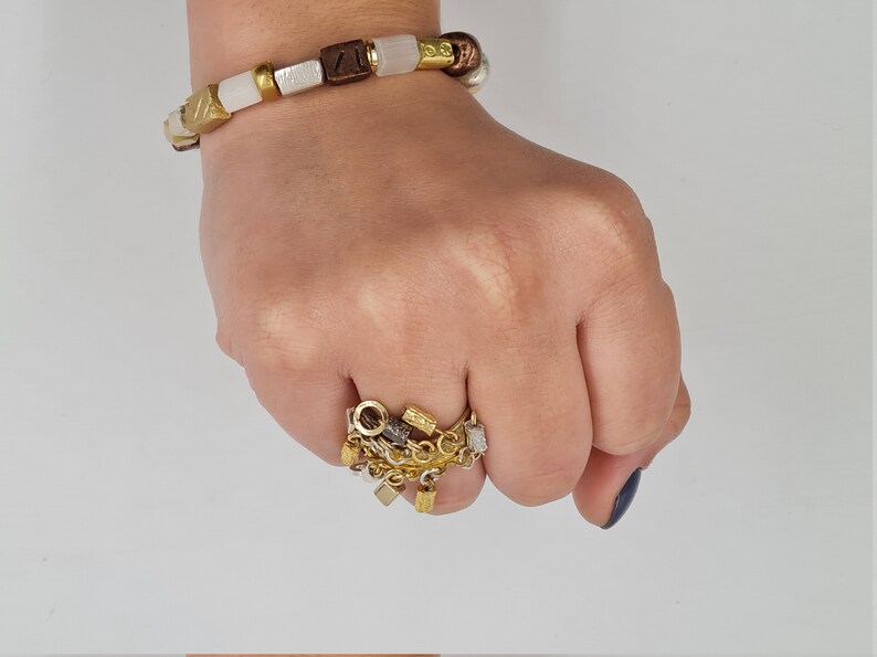 May include: A gold ring with a chain and multiple charms, including a small square, a circle, and a few other shapes. The ring is worn on a person's finger.