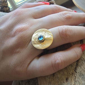 Può includere: Un anello color oro caratterizzato da un grande disco circolare con texture martellata come elemento centrale. Una singola piccola pietra preziosa blu, rotonda e sfaccettata, è incastonata al centro del disco, creando un contrasto con la superficie.