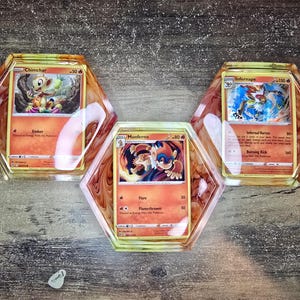 May include: Three Pokémon trading cards set in hexagonal resin coasters. The cards feature Chimchar, Monferno, and Infernape characters. The coasters have a marbled, reddish-brown and white pattern, with a glossy finish.