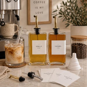 Puede incluir: Dos botellas de jarabe de vidrio rectangulares con vertedores dorados y etiquetas blancas que dicen Vanilla Syrup y Caramel Syrup. Incluye un embudo de silicona blanco, dos vertedores adicionales y un paquete de etiquetas adhesivas extra.