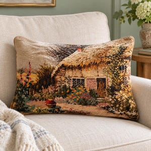 May include: A rectangular throw pillow featuring a woven tapestry design of a cottage with a thatched roof, surrounded by a garden with colorful flowers, greenery, and a path, all rendered in shades of beige, brown, green, and pink.