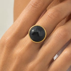 May include: A gold ring featuring a large, smooth, circular black stone set in a simple gold bezel. The ring has a polished finish and a minimalist design, showcasing a deep black cabochon stone centred within a thin, bright gold metal band.