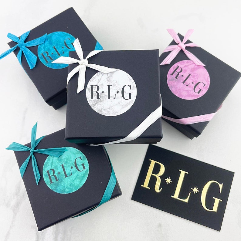 May include: Four black gift boxes with white ribbon and a black card with gold lettering that reads "R*LG". Each box has a different colored circle with the same text in white lettering. The colors are teal, light blue, silver, and pink.