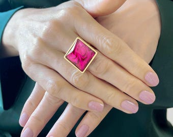 May include: A gold-toned ring featuring a large, square-shaped setting with a vibrant magenta and dark pink marbled resin inlay. The geometric design is centered on the finger, showcasing a smooth, polished metal frame around the colorful stone.