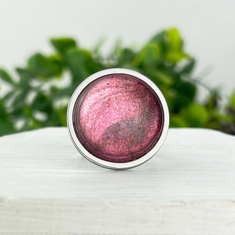May include: A round, silver-rimmed charm with a deep pink, glittery resin insert. The resin has a swirling, marbled effect.