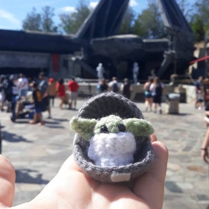 May include: A small crocheted plush toy of a green alien character from the Star Wars franchise, sitting in a gray crocheted cradle. The toy is being held in a person's hand.