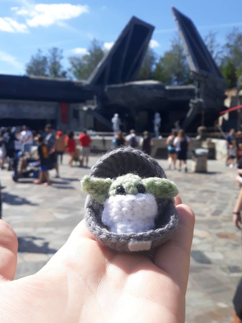 May include: A small crocheted plush toy of a green alien character from the Star Wars franchise, sitting in a gray crocheted cradle. The toy is being held in a person's hand.