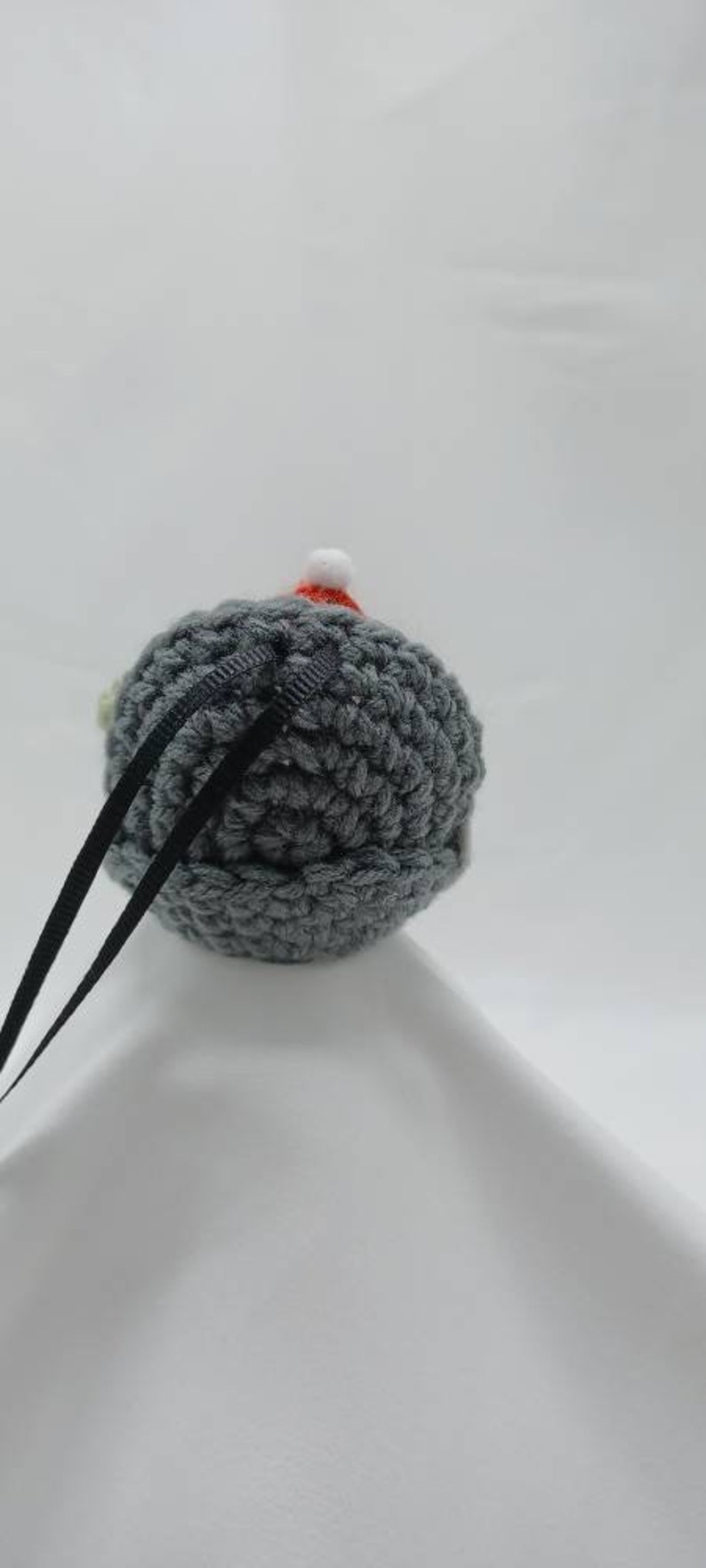May include: A gray crocheted ornament with a red and white crocheted Santa hat. The ornament has a black ribbon attached to it.