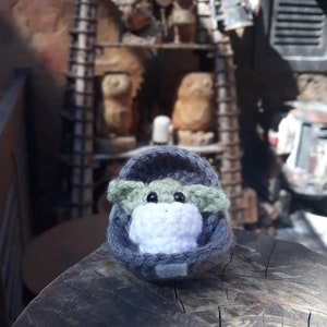 May include: A small green crocheted character with large eyes, wearing a gray crocheted hooded robe, sitting on a wooden surface.